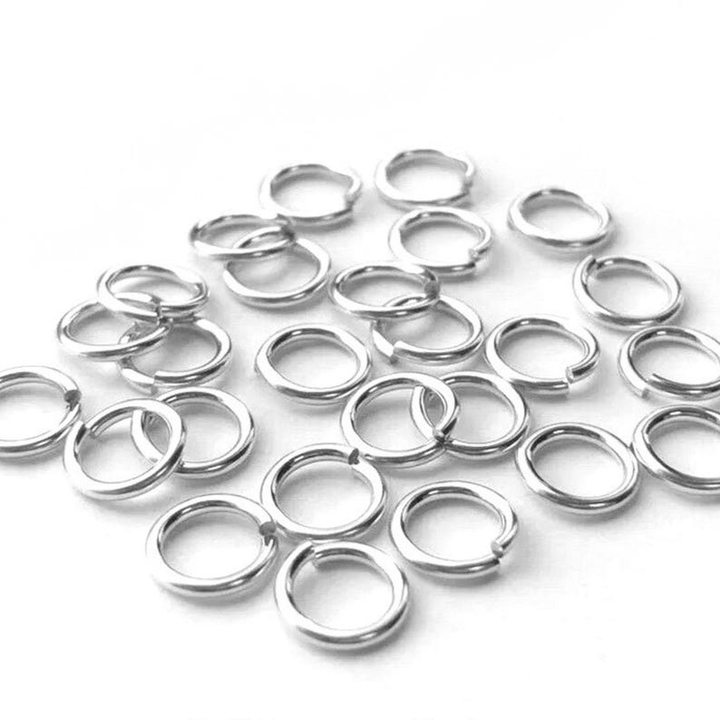 May include: A collection of small, silver-colored jump rings scattered on a white surface. The rings are circular with a small opening, designed for jewelry making or crafts. The rings are made of a shiny, metallic material.