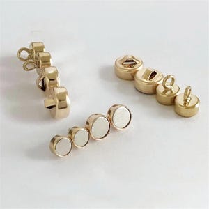 May include: A collection of gold-colored magnetic clasps and buttons. The clasps have a loop for attaching to fabric, while the buttons have a flat back with a magnet. The clasps and buttons are in various sizes and shapes.