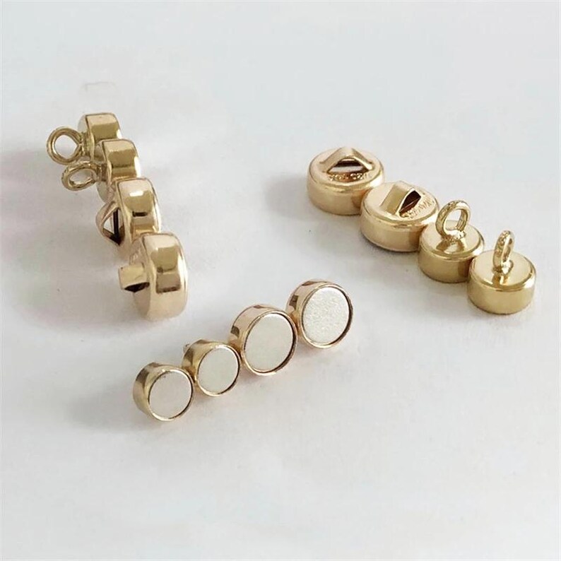 May include: A collection of gold-colored magnetic clasps and buttons. The clasps have a loop for attaching to fabric, while the buttons have a flat back with a magnet. The clasps and buttons are in various sizes and shapes.