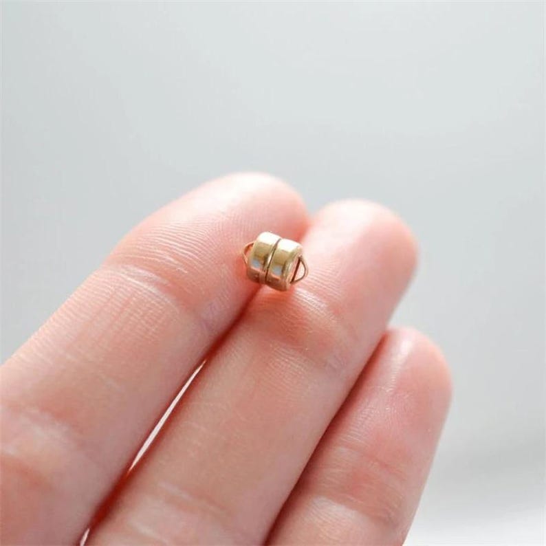 May include: A small, gold-colored magnetic clasp, composed of two cylindrical pieces held together. The clasp is designed for jewelry making or similar crafts. The image is a close-up, showing the clasp held between fingers against a soft, neutral background.