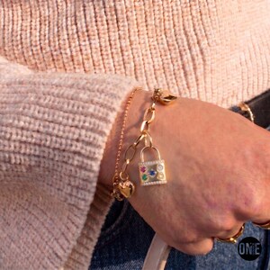 May include: A gold-toned chain bracelet featuring a padlock charm adorned with small, colorful gemstones including pink, blue, green, and white stones, alongside a small heart charm, worn on a wrist with a light-colored, textured knit sweater.