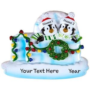 May include: Personalized Christmas ornament featuring two penguins peeking out of an igloo decorated with a wreath and Christmas lights. The ornament has the text "Your Text Here" and "Year" on the bottom.
