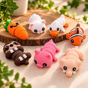 Op de afbeelding: Een verzameling van zeven kleine, beweegbare dieren- en groentefiguren, waaronder een oranje wortel, een witte kat, een witte eend, een oranje met witte clownvis, een bruine luiaard, een roze varken en een lichtroze olifant met grote oren.
