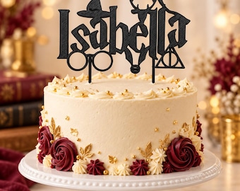 May include: A black cake topper featuring the name Isabella in a gothic font, topped with a witch hat, round glasses, a deer silhouette, and a triangular symbol, placed on a round cake with white frosting, gold sprinkles, and dark red roses.