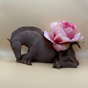 Peut inclure: Une sculpture de cheval en céramique brune avec une rose artificielle rose sur le dos. Le cheval est couché, la tête baissée.