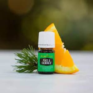 May include: A small, dark brown glass bottle with a white cap, labeled "Peace & Calming II" essential oil blend. The bottle is sitting on a wooden surface with a green pine branch and a slice of orange.