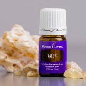 May include: A small, purple bottle of Young Living essential oil labeled "Valor". The bottle is sitting on a white surface with a few pieces of amber-colored resin.
