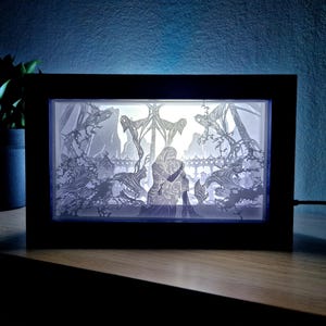 May include: A framed, illuminated paper cut artwork. The piece shows a couple embracing in a detailed, layered scene with trees and architectural elements. The frame is black, and the background is lit with a soft glow.