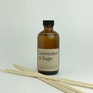 May include: A brown glass bottle with a black lid and a label that reads "Lavender & Sage". The bottle is filled with a clear liquid and is sitting on a white surface. There are wooden sticks in front of the bottle.