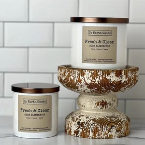 May include: Two white candles with copper lids and brown labels. The labels read "Up North Goods" and "Fresh & Clean Odor Eliminator" with the scent notes "Linen | Ozone | Lemon". The candles are on a distressed white wooden pedestal.