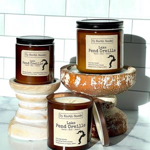 May include: Three brown glass candle jars with black lids. Each jar has a label that reads "Up North Goods" and "Lake Pend Oreille". The label also includes the scent "Citrus | Mint | Jasmine" and a small outline of a lake. The candles are on a white wooden pedestal.
