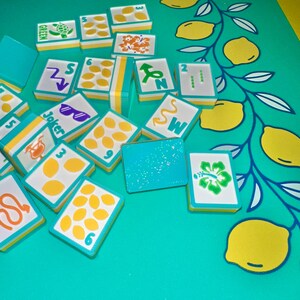 May include: A collection of colorful Mahjong tiles with various symbols and numbers, including lemons, flowers, and sunglasses. The tiles are arranged on a turquoise surface with a lemon-themed design. The tiles have a white background with yellow and turquoise borders.