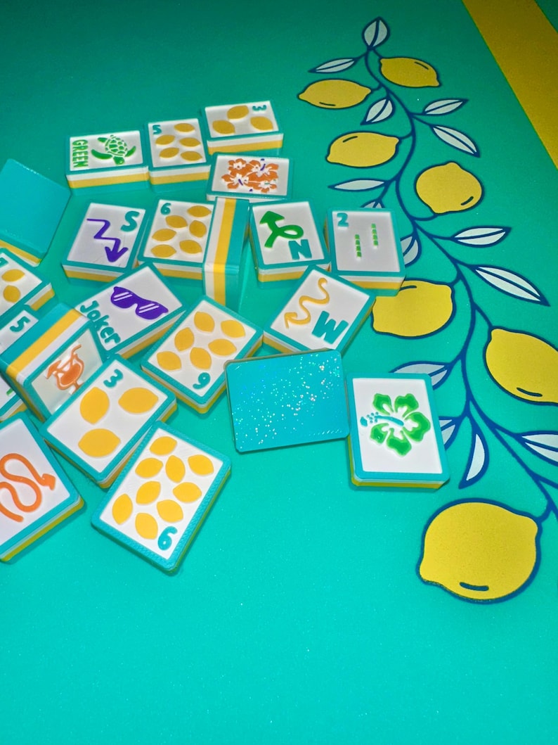 May include: A collection of colorful Mahjong tiles with various symbols and numbers, including lemons, flowers, and sunglasses. The tiles are arranged on a turquoise surface with a lemon-themed design. The tiles have a white background with yellow and turquoise borders.
