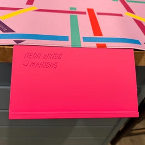 May include: A bright pink rectangular mahjong rack with the embossed text NEON WINDS and a heart symbol followed by MAHJONG. Positioned above it is a light pink game mat featuring a grid of intersecting lines in red, yellow, blue, and green.