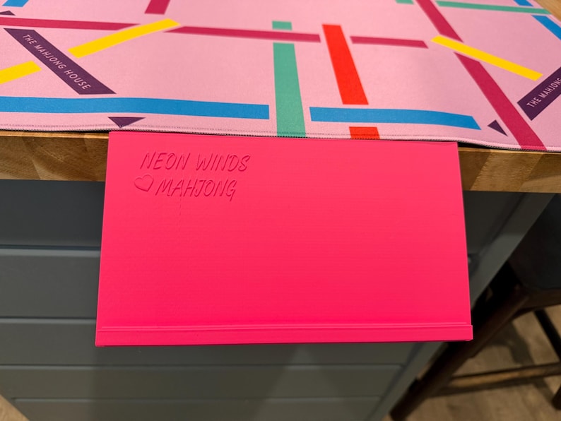 May include: A bright pink rectangular mahjong rack with the embossed text NEON WINDS and a heart symbol followed by MAHJONG. Positioned above it is a light pink game mat featuring a grid of intersecting lines in red, yellow, blue, and green.