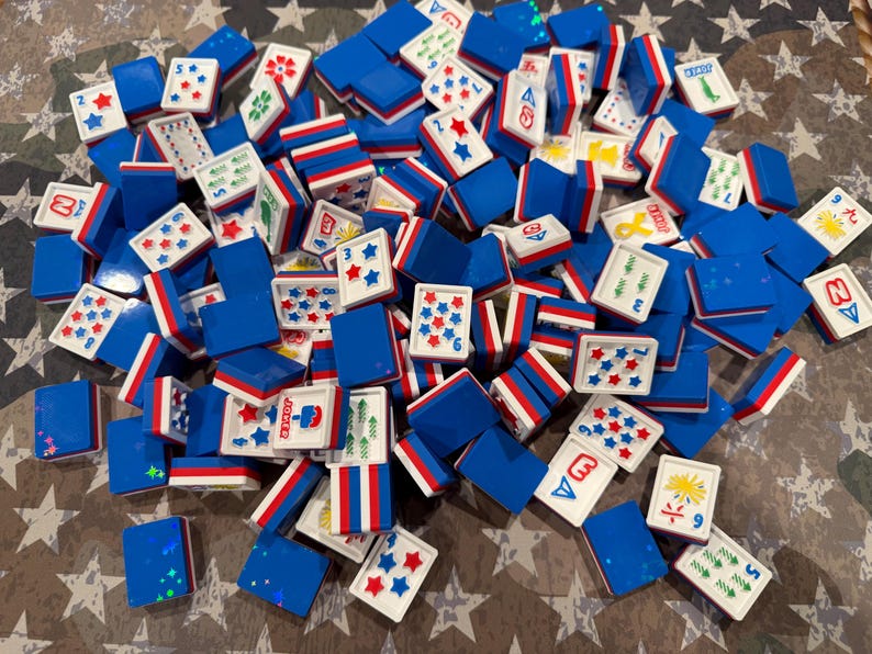 May include: A collection of blue, white, and red Mahjong tiles scattered across a surface. The tiles feature various symbols, numbers, and star patterns. The tiles are rectangular with a blue base and white tops with red and blue stripes.