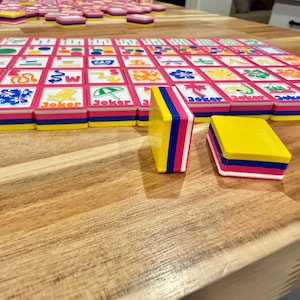 May include: A colorful Mahjong set with pink, blue, and yellow tiles. The tiles are arranged on a wooden surface, with some tiles stacked and others laid out in a grid. The word "Joker" is printed on some tiles.