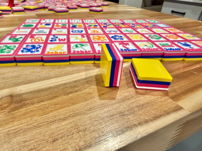 May include: A colorful Mahjong set with pink, blue, and yellow tiles. The tiles are arranged on a wooden surface, with some tiles stacked and others laid out in a grid. The word "Joker" is printed on some tiles.