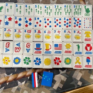 May include: A complete set of white Mahjong tiles with various symbols, numbers, and characters in red, blue, and green. Includes joker tiles and two small blue and red tile holders. The game set is displayed on a patterned surface.