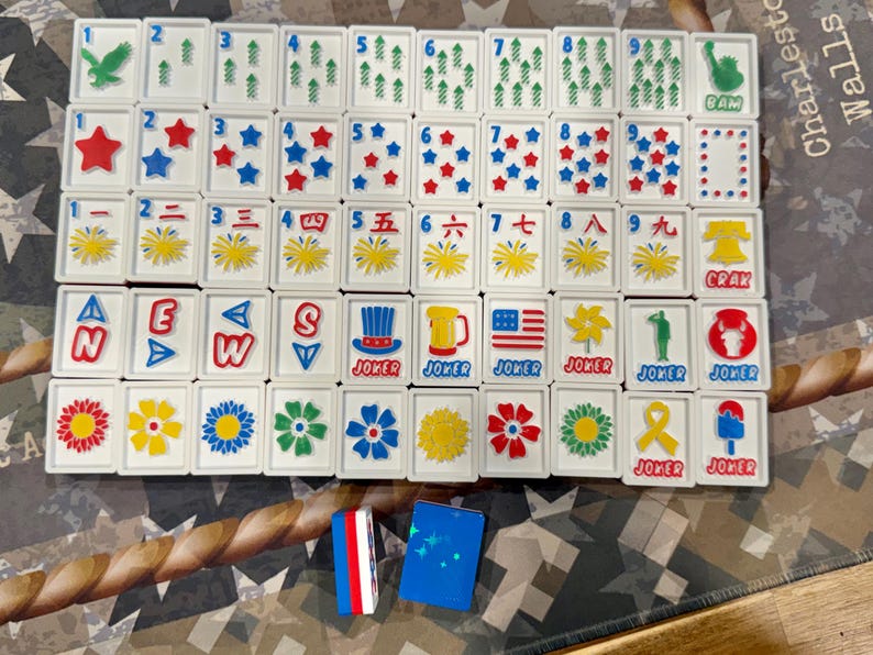 May include: A complete set of white Mahjong tiles with various symbols, numbers, and characters in red, blue, and green. Includes joker tiles and two small blue and red tile holders. The game set is displayed on a patterned surface.