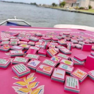 May include: A pink Mahjong set with many tiles on a matching pink surface. The tiles feature various symbols and characters, with pink, white, and blue accents. The background shows water and a boat.