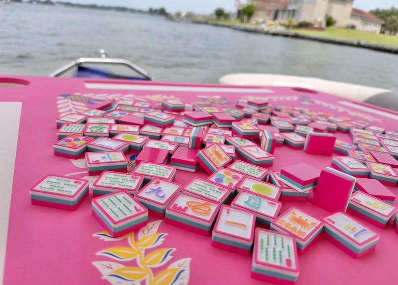 May include: A pink Mahjong set with many tiles on a matching pink surface. The tiles feature various symbols and characters, with pink, white, and blue accents. The background shows water and a boat.