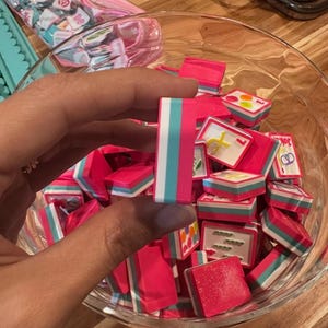 May include: A clear glass bowl filled with small, pink, rectangular objects with white, teal, and pink stripes. One object is held by a hand. Other objects in the bowl have various designs. A pink bag with small, square objects is in the background.