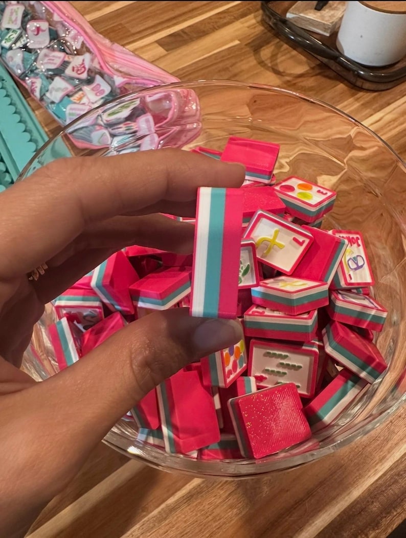 May include: A clear glass bowl filled with small, pink, rectangular objects with white, teal, and pink stripes. One object is held by a hand. Other objects in the bowl have various designs. A pink bag with small, square objects is in the background.