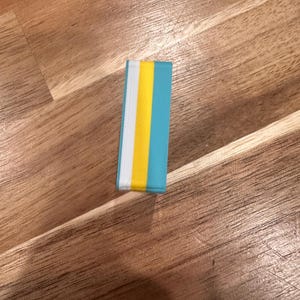 May include: A rectangular eraser with a teal, yellow, white, and teal striped design. The eraser is lying on a wooden surface.