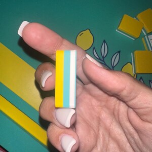May include: A rectangular eraser with yellow, light blue, and white stripes. The eraser is held in a hand with manicured nails. The background features a teal surface with yellow and white accents, and lemon illustrations.