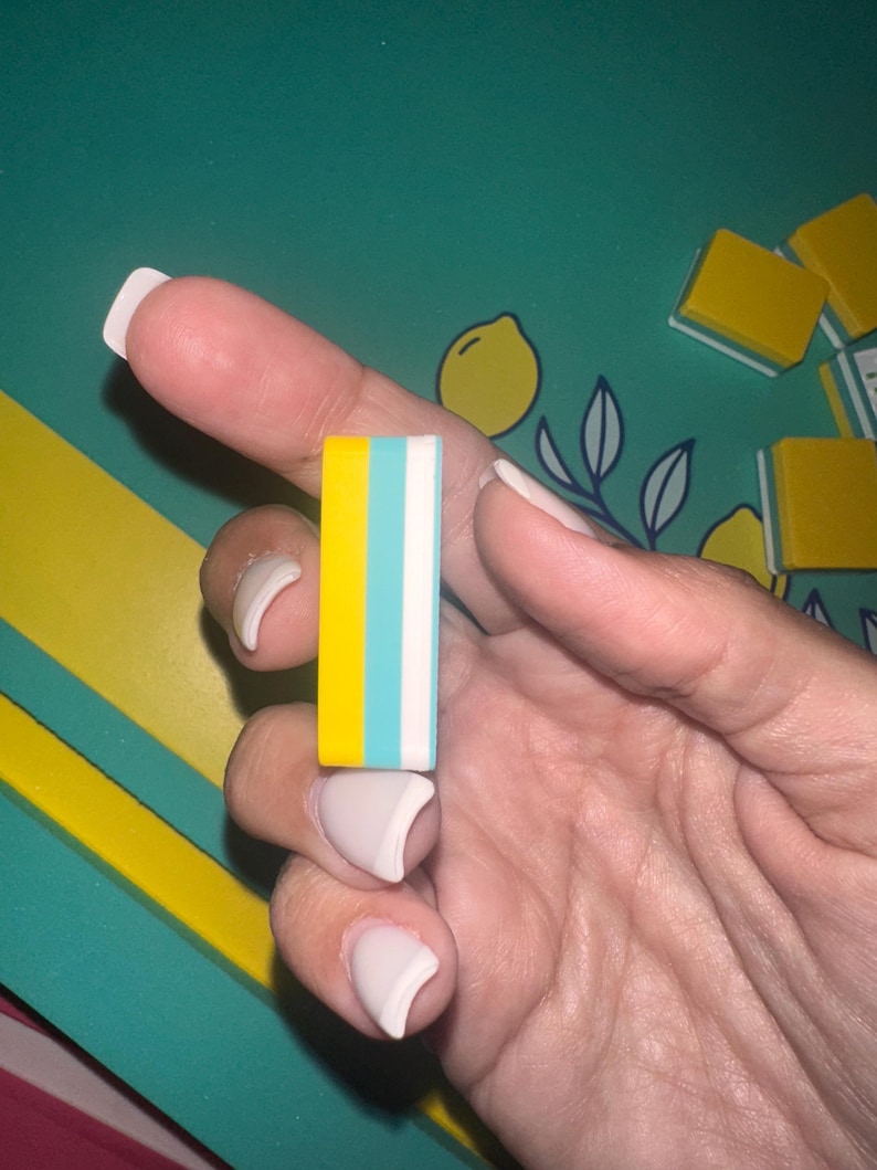 May include: A rectangular eraser with yellow, light blue, and white stripes. The eraser is held in a hand with manicured nails. The background features a teal surface with yellow and white accents, and lemon illustrations.