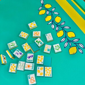 May include: A teal game board with yellow and white lemon branch design. Scattered across the board are small rectangular game tiles with various symbols, numbers, and the word "Joker". The tiles have teal backs and white faces with yellow accents.