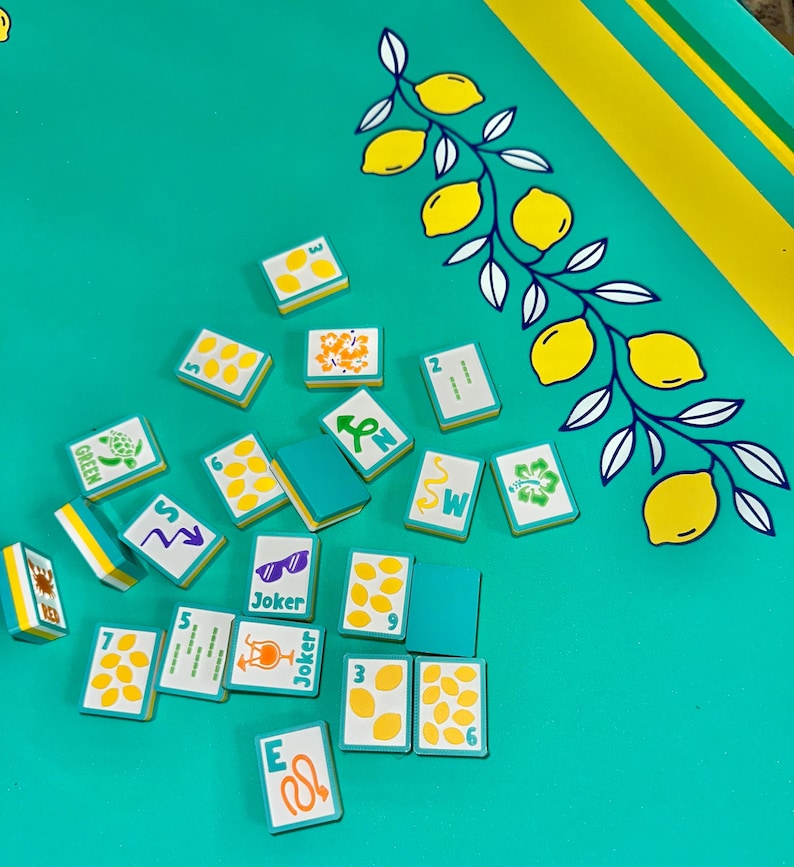 May include: A teal game board with yellow and white lemon branch design. Scattered across the board are small rectangular game tiles with various symbols, numbers, and the word "Joker". The tiles have teal backs and white faces with yellow accents.