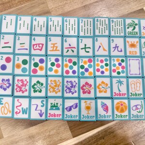 May include: A full set of Mahjong tiles with a turquoise border. The tiles display various symbols, numbers, and characters in green, orange, and purple. Some tiles have the words "Joker", "Green", and "Red".