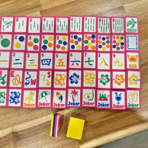 May include: A complete Mahjong set with pink-framed tiles arranged on a wooden surface. The tiles feature various symbols, numbers, and characters in multiple colors. Two additional tiles are placed below the main set. The word "Joker" is printed on some tiles.