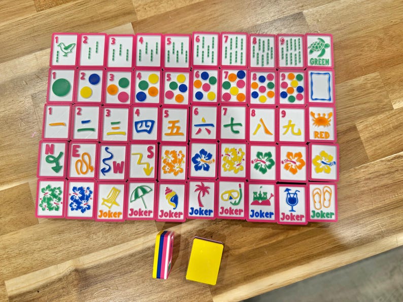 May include: A complete Mahjong set with pink-framed tiles arranged on a wooden surface. The tiles feature various symbols, numbers, and characters in multiple colors. Two additional tiles are placed below the main set. The word "Joker" is printed on some tiles.