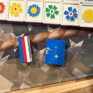 May include: A collection of colorful Mahjong tiles with flower designs and two blue and white game pieces. The tiles feature red, yellow, blue, and green flowers. The game pieces are blue with star accents and red, white, and blue stripes.