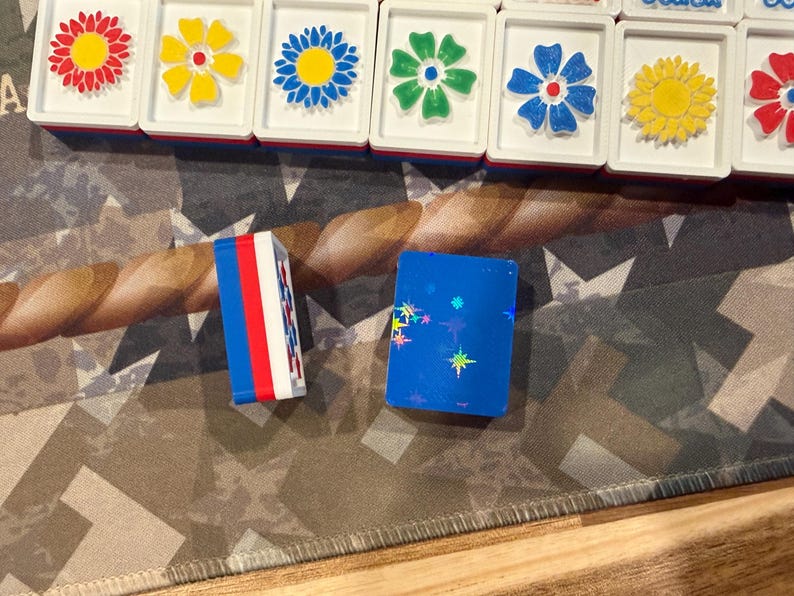 May include: A collection of colorful Mahjong tiles with flower designs and two blue and white game pieces. The tiles feature red, yellow, blue, and green flowers. The game pieces are blue with star accents and red, white, and blue stripes.