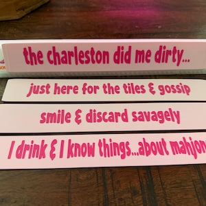 May include: Four rectangular light pink markers with dark pink text. The text reads: the charleston did me dirty..., just here for the tiles & gossip, smile & discard savagely, and I drink & I know things...about mahjong. Each marker is flat.