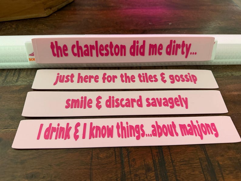 May include: Four rectangular light pink markers with dark pink text. The text reads: the charleston did me dirty..., just here for the tiles & gossip, smile & discard savagely, and I drink & I know things...about mahjong. Each marker is flat.