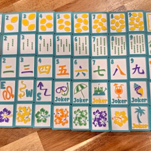 May include: A complete set of Mahjong tiles with a tropical theme. The tiles feature various symbols, numbers, and characters in vibrant colors, including green, red, and yellow. The set includes "Joker" tiles and is arranged on a wooden surface.
