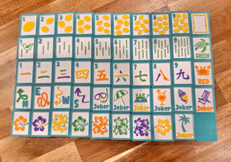 May include: A complete set of Mahjong tiles with a tropical theme. The tiles feature various symbols, numbers, and characters in vibrant colors, including green, red, and yellow. The set includes "Joker" tiles and is arranged on a wooden surface.