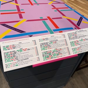 May include: A rectangular light purple Mahjong mat featuring a geometric pattern of intersecting red, blue, yellow, and teal lines with the text THE MAHJONG HOUSE, accompanied by a white Mahjong score card with red, green, and black printed text.