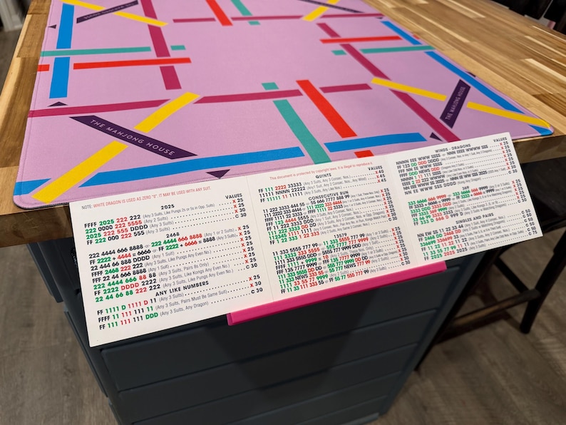 May include: A rectangular light purple Mahjong mat featuring a geometric pattern of intersecting red, blue, yellow, and teal lines with the text THE MAHJONG HOUSE, accompanied by a white Mahjong score card with red, green, and black printed text.