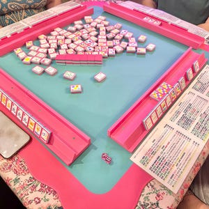 May include: A pink Mahjong set with a teal playing surface. The set includes tiles, racks, and dice. A smartphone and a printed sheet with text are also visible on the table. The table is set on a floral patterned tablecloth.