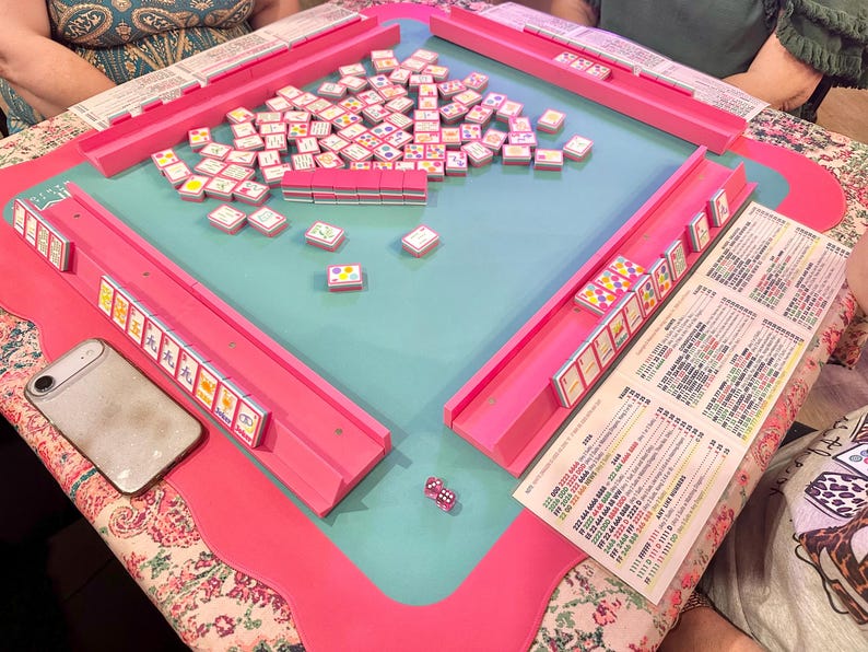 May include: A pink Mahjong set with a teal playing surface. The set includes tiles, racks, and dice. A smartphone and a printed sheet with text are also visible on the table. The table is set on a floral patterned tablecloth.