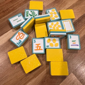 May include: A collection of yellow and white game tiles with blue accents. Some tiles feature numbers, symbols, and illustrations, including a turtle. The tiles are scattered on a wooden surface.