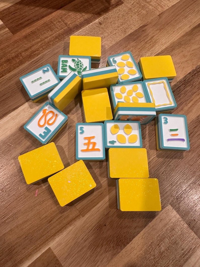 May include: A collection of yellow and white game tiles with blue accents. Some tiles feature numbers, symbols, and illustrations, including a turtle. The tiles are scattered on a wooden surface.