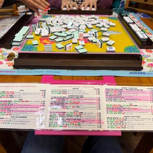 May include: A Mah Jongg game set featuring a yellow playing mat with a floral border, dark brown tile racks, and a collection of light green and white tiles. A tri-fold Mah Jongg score card is displayed on a bright pink stand in the foreground.