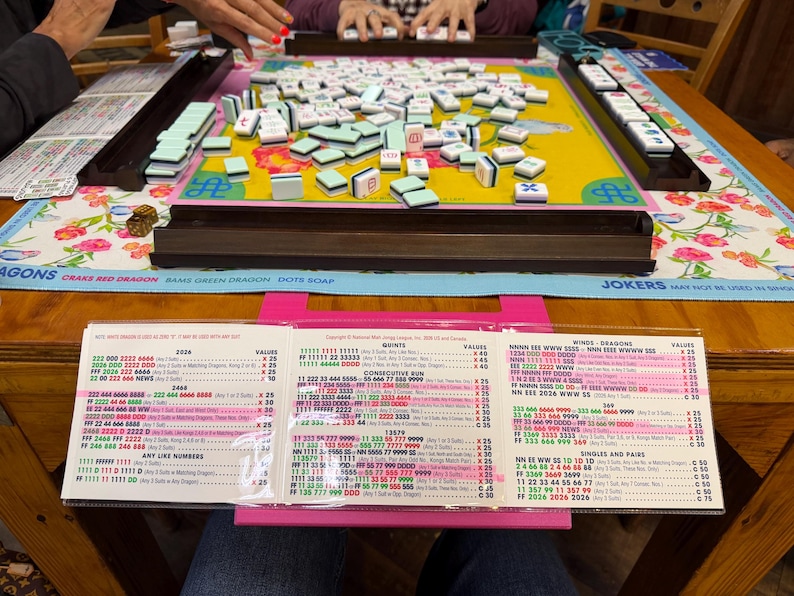 May include: A Mah Jongg game set featuring a yellow playing mat with a floral border, dark brown tile racks, and a collection of light green and white tiles. A tri-fold Mah Jongg score card is displayed on a bright pink stand in the foreground.